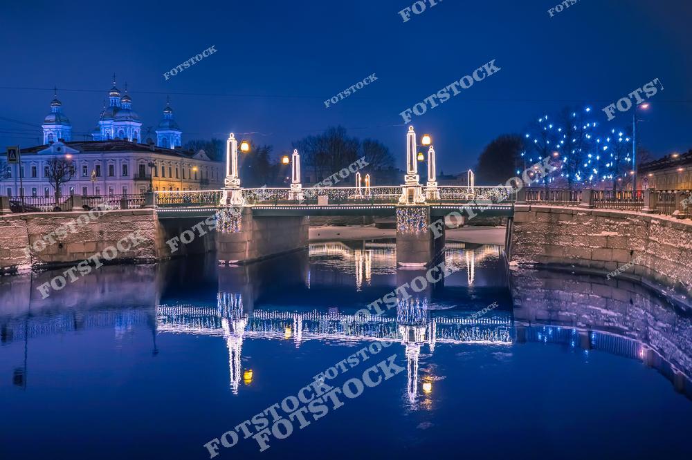Фотография вечернего Санкт-Петербург, Россия, в период рождественских и новогодних праздников. На изображении запечатлены реки и мосты Санкт-Петербурга, украшенные праздничной иллюминацией. Фотоснимок показывает декорированный мост и набережную Крюков канал в вечернее время. Фото передаёт атмосферу Нового года и Рождества в городе: огни гирлянд отражаются в воде, создавая уютный и праздничный облик зимнего Петербурга. Иллюстрация может использоваться как изображение, картинка или слайд, посвящённые зимним праздникам, мостам и рекам Санкт-Петербурга.